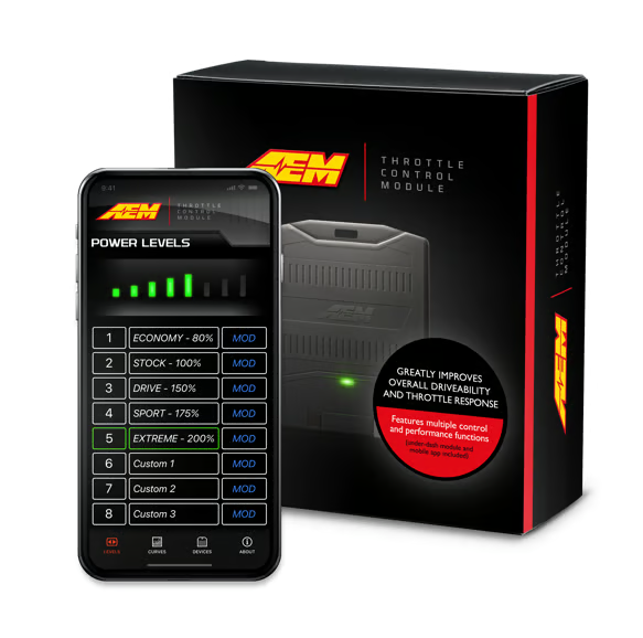 A black AEM Throttle Control Module shown in front of its retail box, with a smartphone displaying the AEM app power-level interface. The box is black with orange and red branding and text, and the phone screen is black with green and white interface elements.